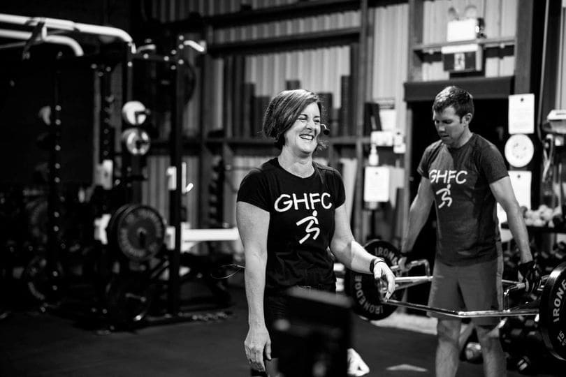 Two people exercising in a gym while wearing "GHFC" t-shirts, with one smiling woman holding a barbell in the foreground and a man lifting weights in the background.